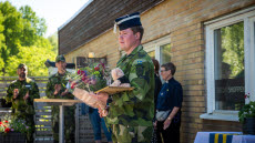 Soldat står i förgrunden med blommor och en nalle. I bakgrunden står två militärer och klappar händerna samt en civilklädd kvinna. 