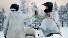Två soldater i vinteruniform och skoterhjälmar gör en high five. Mellan dem står en skoter. Landskapet är snöklätt och träd syns i bakgrunden. 