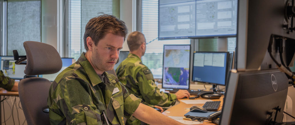 Två män i uniform sitter vid datorskärmar och kartor