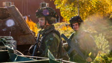 Två manliga soldater i grön uniform och hjälmar bärandes vapen syns på bilden. Den ena ser allvarlig ut och den andra ler. I bakgrunden syns en röd byggnad samt träd. 