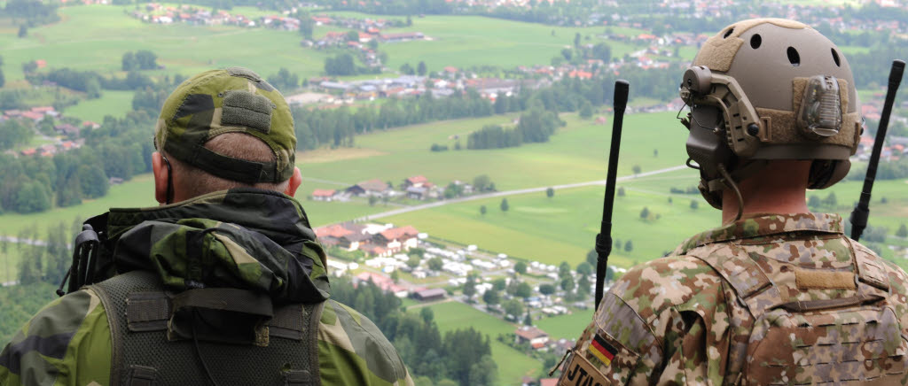 Svensk JTAC och tysk JTAC koordinerar tillsammans ett flyganfall mot en fientlig styrka som anfaller ett mindre samhälle.