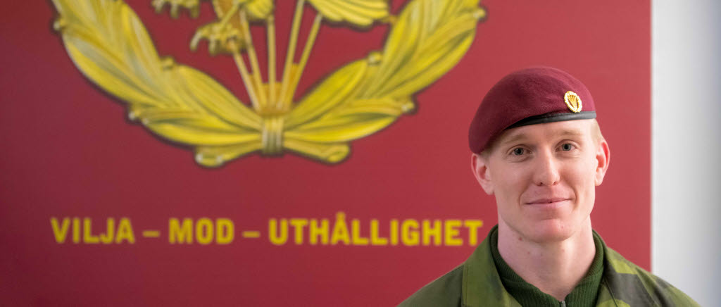 Anton iklädd fallskärmsjägarnas vinröda basker. I bakgrunden ses fallskärmsjägarnas ledord: Vilja - Mod - Uthållighet.