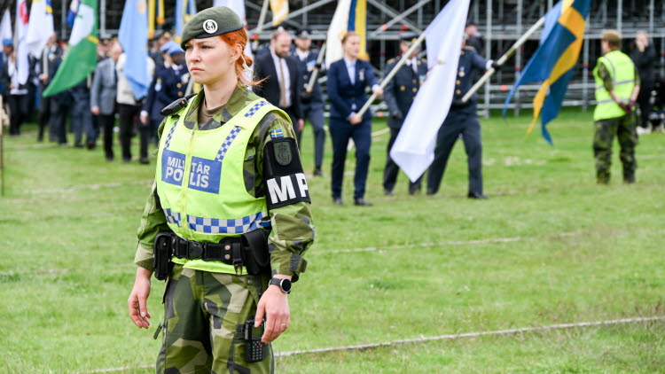 Kvinnlig militärpolis står och tittar framåt. Bakom henne går människor med olika flaggor. 