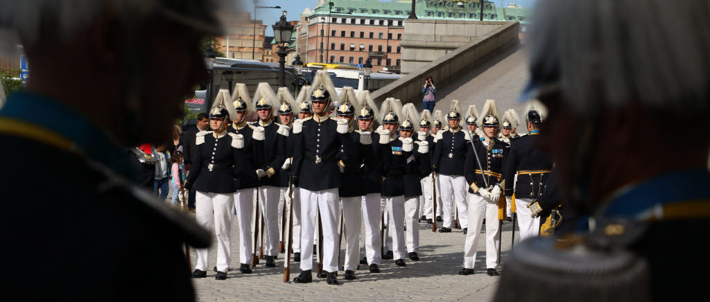 Soldater med hjälmar med ceremoniella plymer, uniformer med axelklaffar och vita byxor står uppradade. 