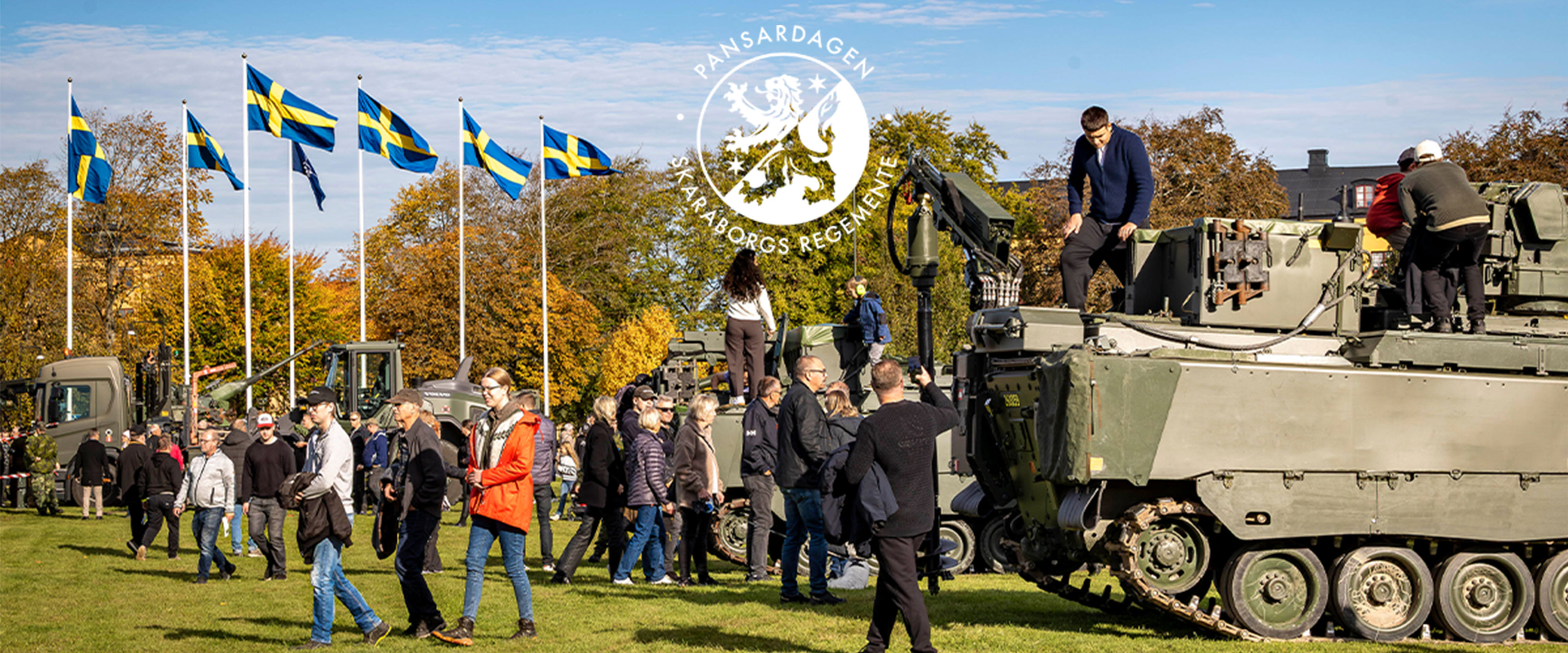 Personer som tittar på militärfordon på en öppen yta. Flaggor i bakgrunden.