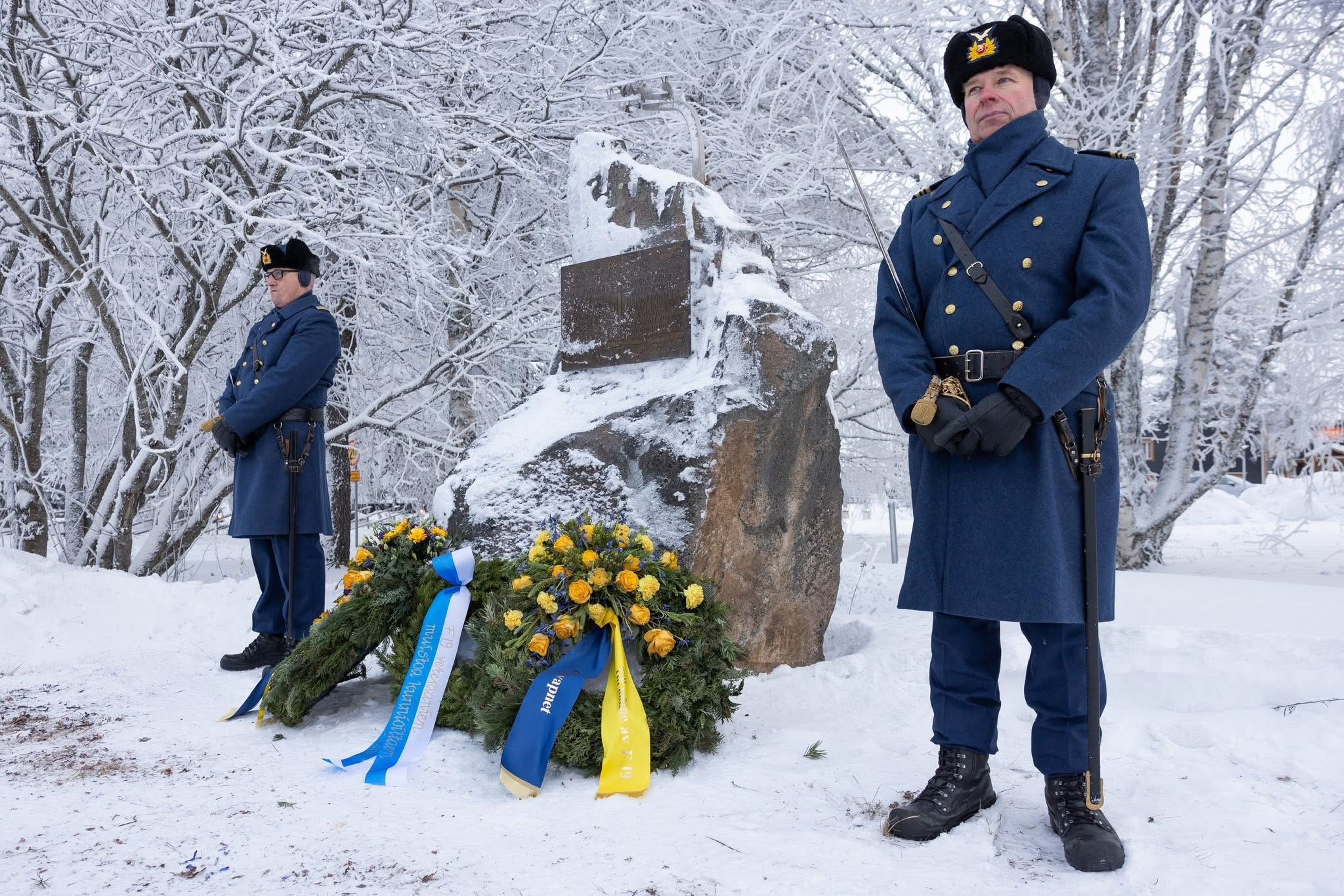 Två personer i uniform står bredvid en minnessten där det ligger blomkransar.