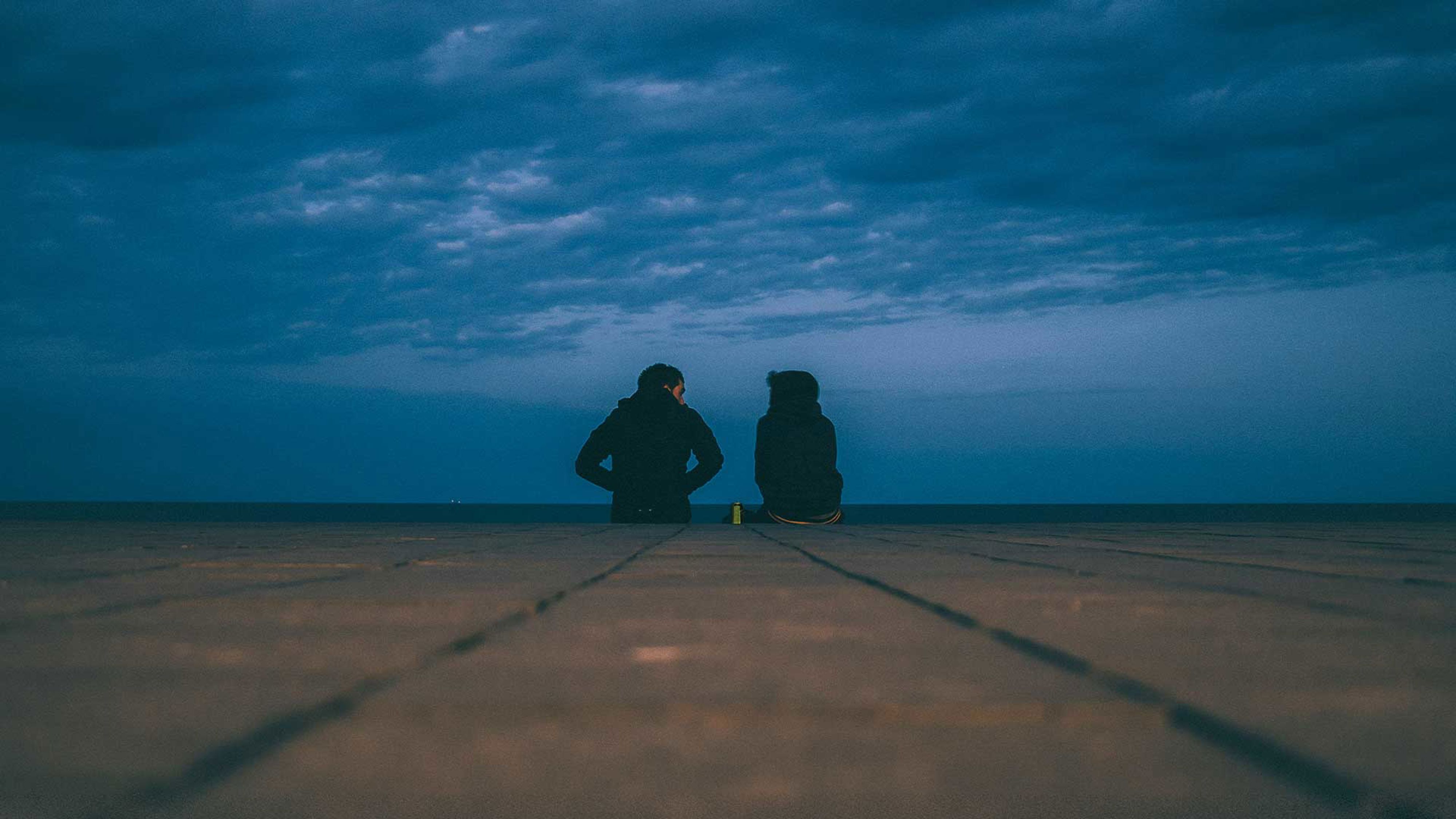 två personer som sitter med ryggen mot fotografen i mörka kläder under mörk himmel