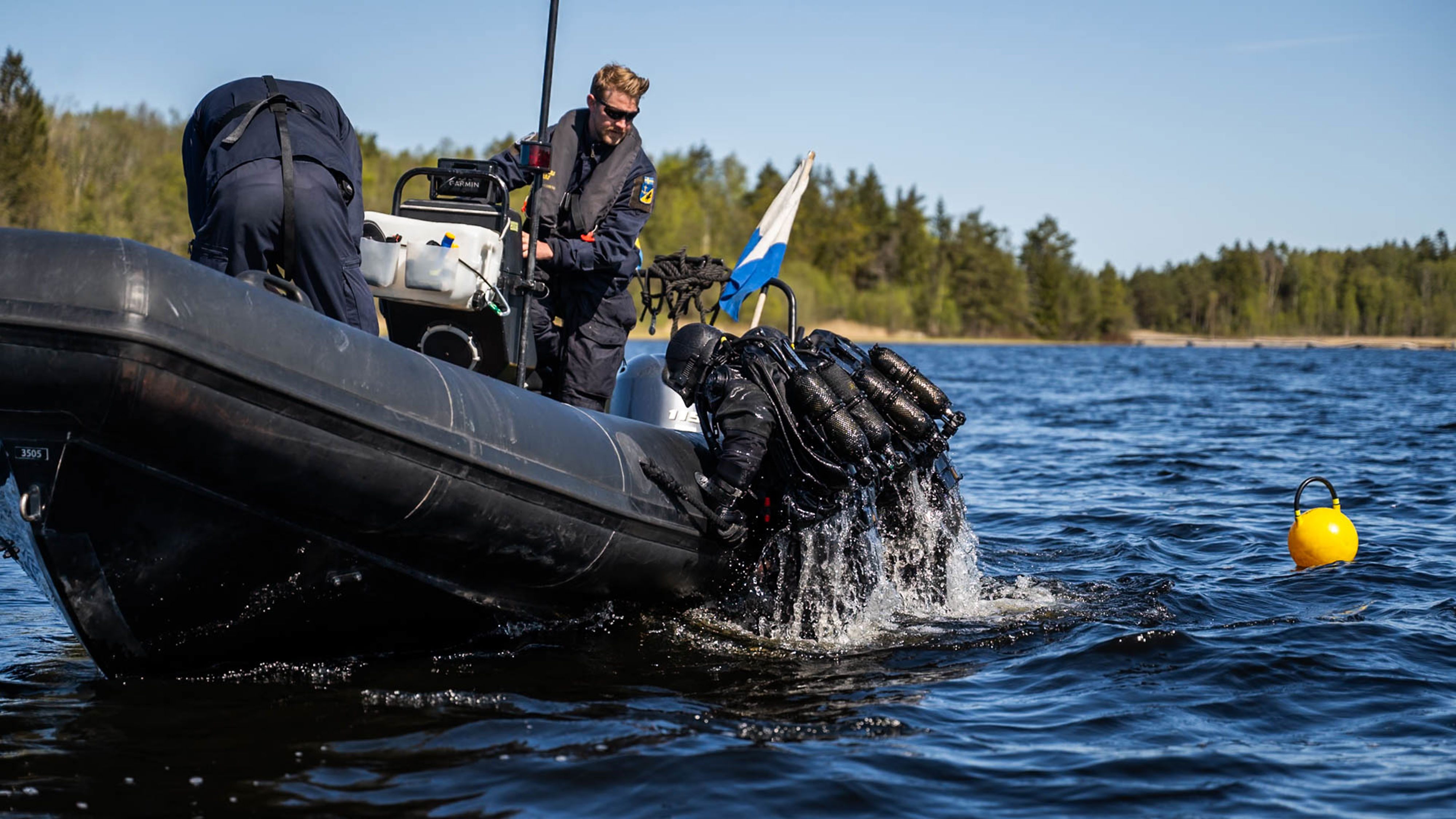 Två dykare med svart dykutrustning häver sig upp i en svart RHIB-båt på en klarblå sjö under en blå himmel. På vattenytan guppar en gul boj. I båten arbetar två personer i marinens blå fältuniform och flytväst. Båten har flagga Adam hissad, som visar att det finns dykare i vattnet.