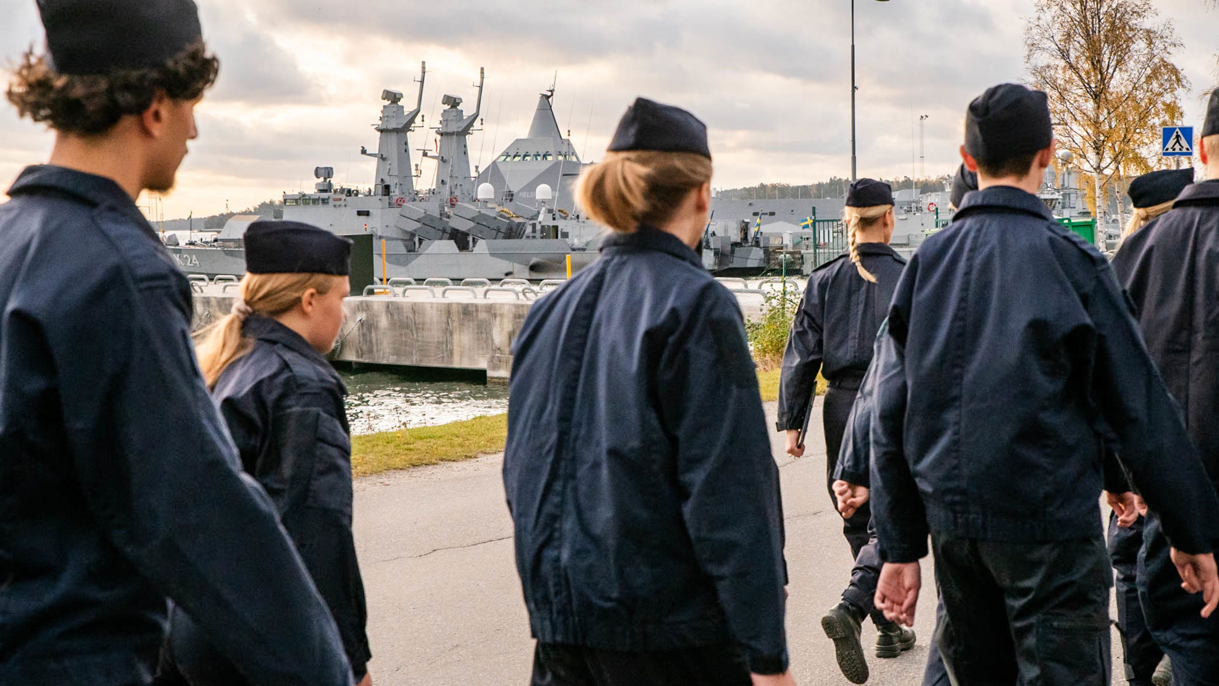 En grupp ungdomar i marinens mörkblå uniform går längs en kaj framför svenska marinens fartyg i en örlogshamn.