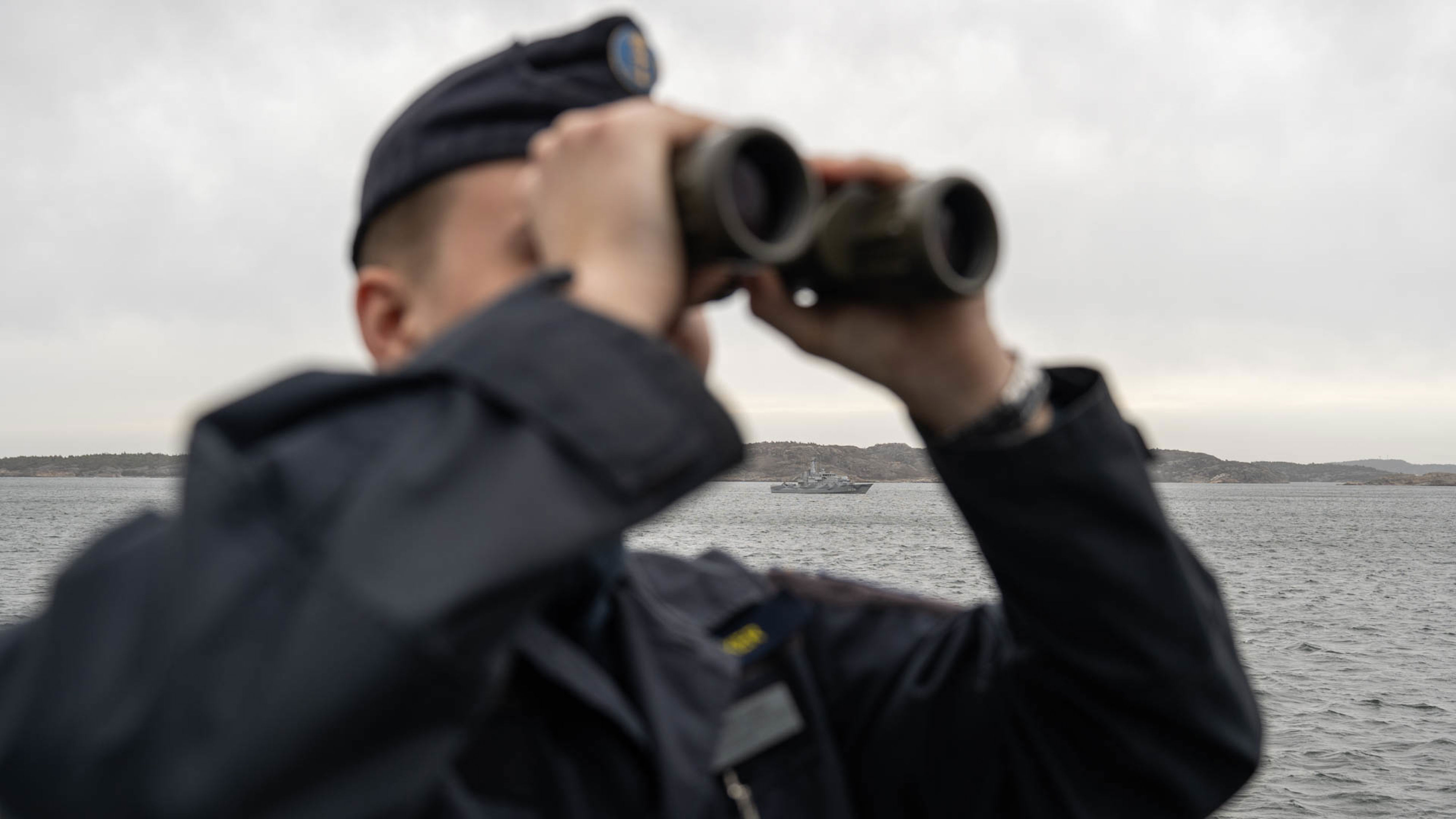 Sjöman med marinens blå uniform spanar med kikare över ett grått hav. Ett grått minröjningsfartyg syns längre bort.