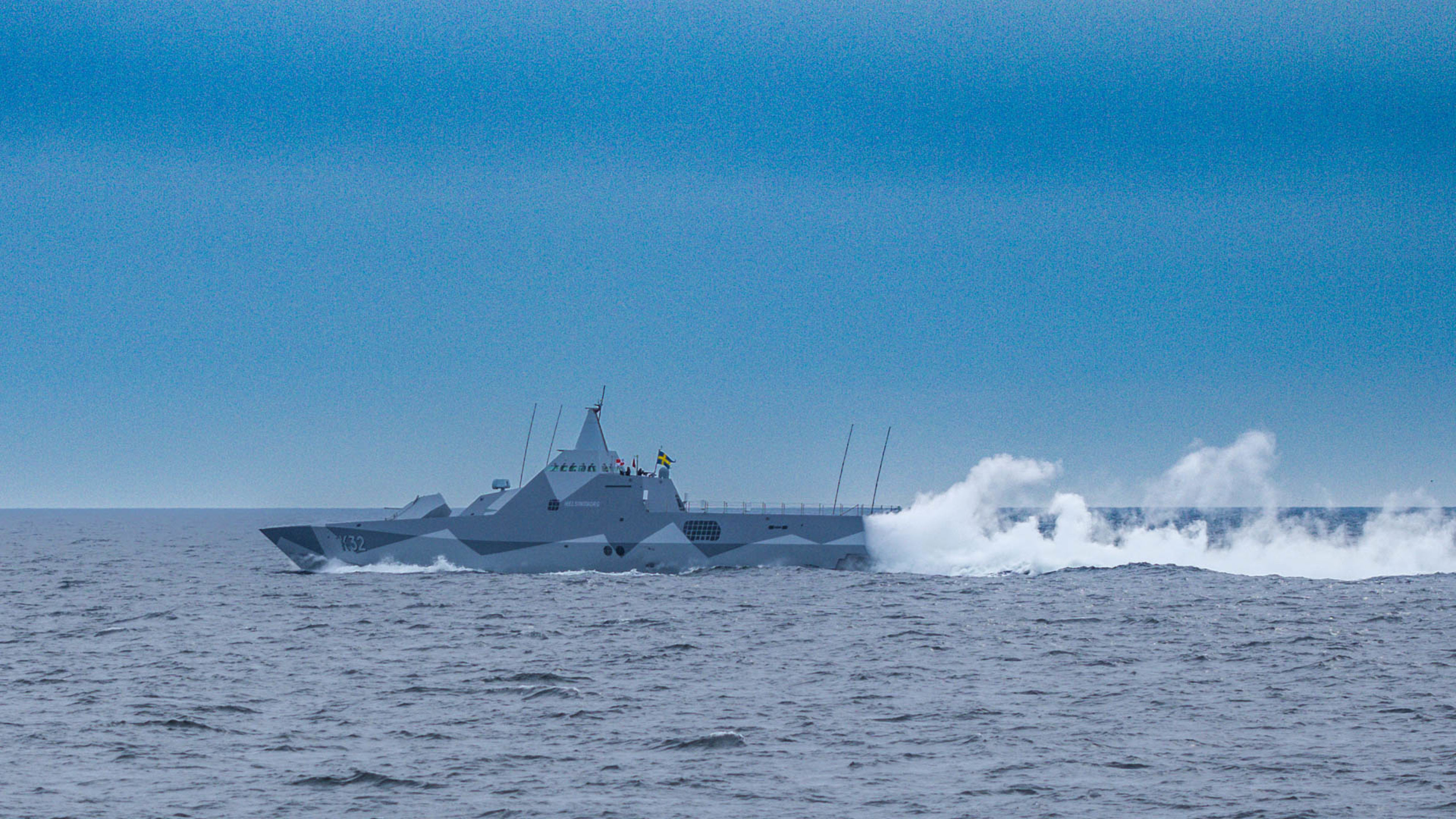 En grått örlogsfartyg med spetsiga och svenska flagga forsar fram över vattnet med vit rök bakom.