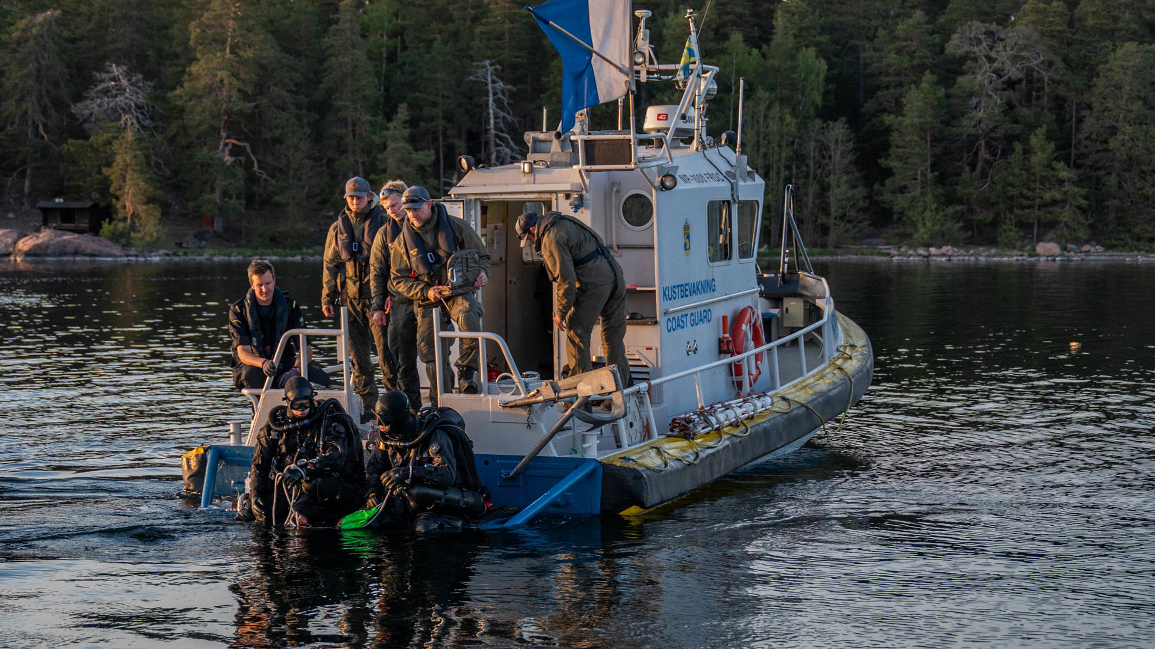 Marinens röjdykare förbereder sig på att dyka från en av Kustbevakningens mindre båtar. Dykare sitter med benen i vatten bakom båten. Militärer och kustbevakare befinner sig på båtens akterdäck. Kvällsljuset lyser upp scenen från sidan. En skogsklädd strandkant syns i bakgrunden.