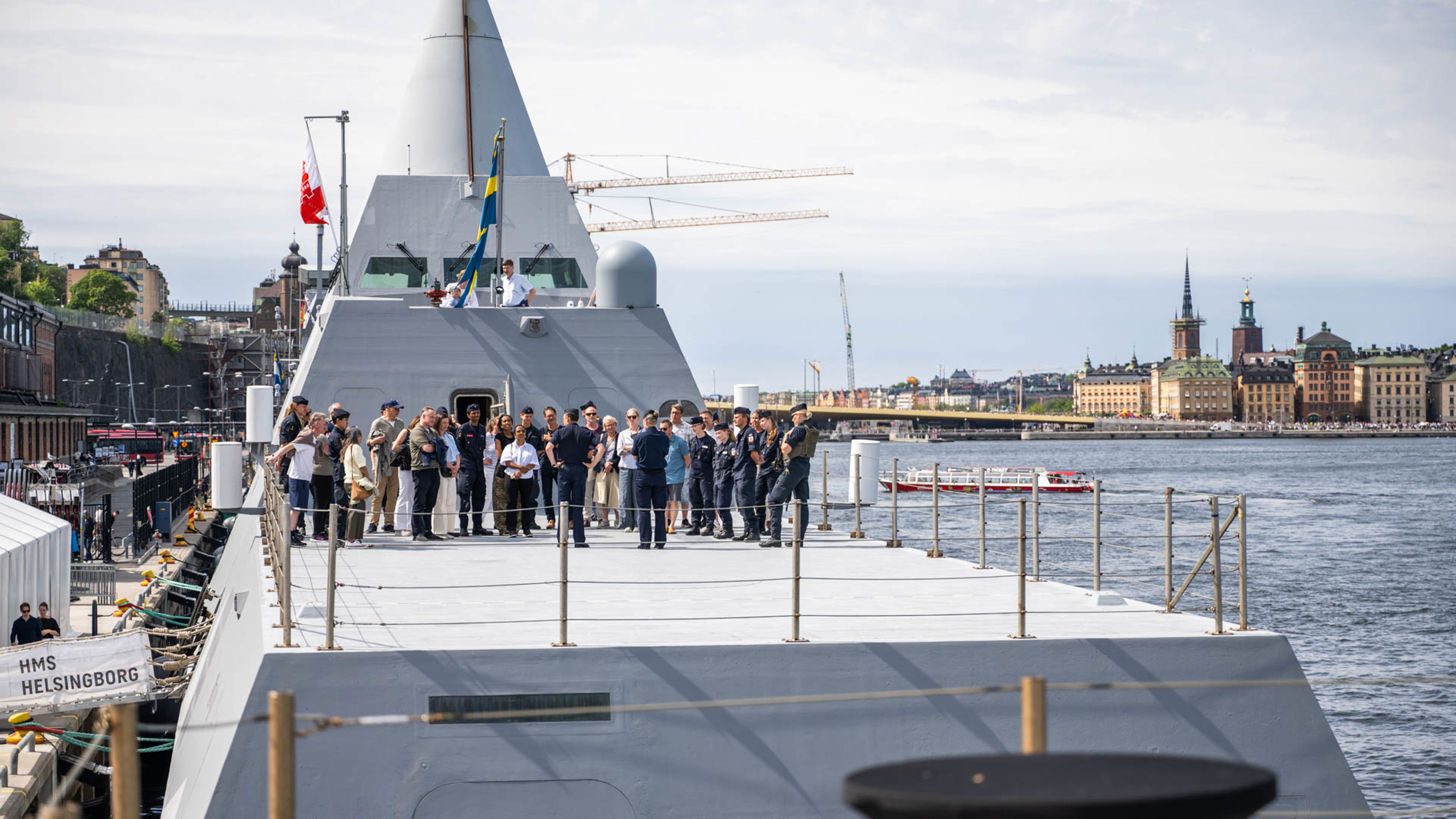 En grå Visbykorvett ligger förtöjd med Saltsjön och Gamla stan i Stockholm i bakgrunden. Många människor med och utan uniform har samlats på akterdäck.