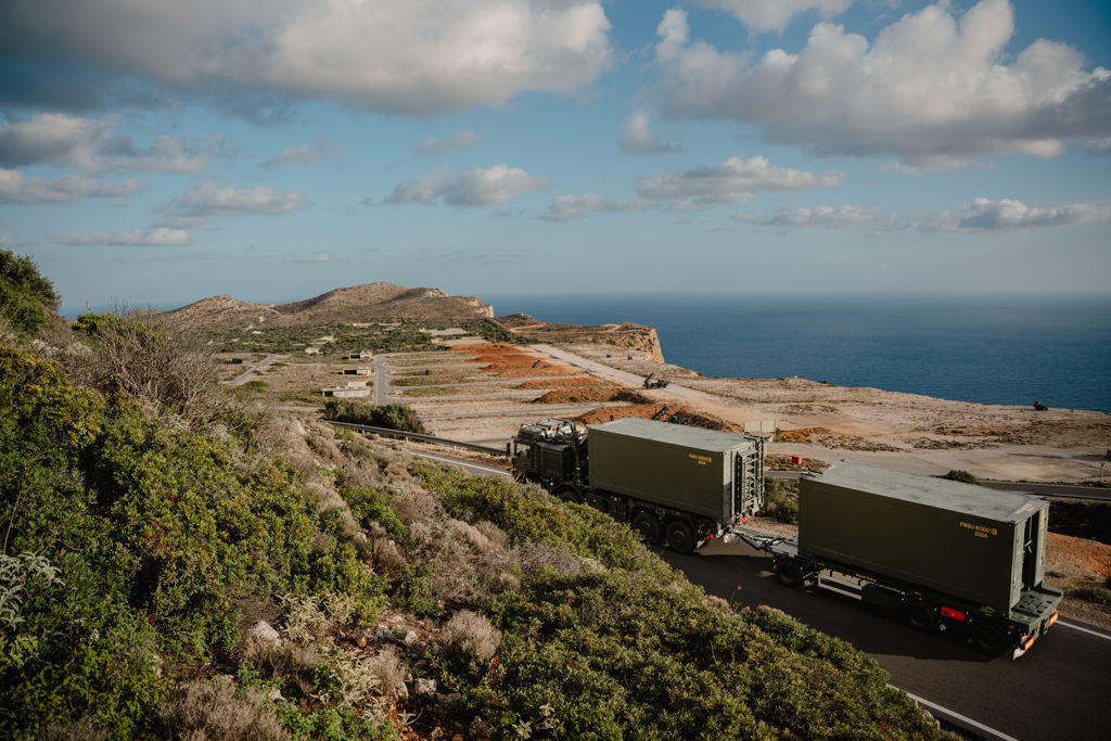 Militära fordon kör på en smal väg intill havet i ett medelhavslandskap