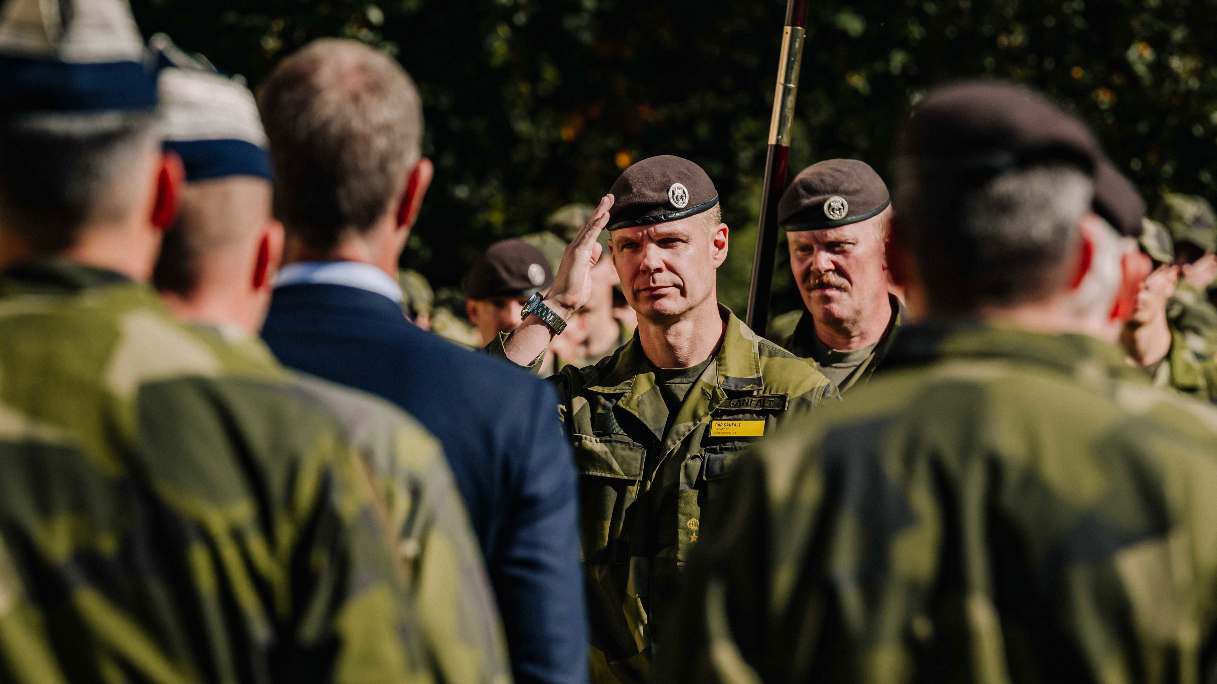I förgrunden syns flera ryggar på grönklädda militärer. I fokus i bilden är en annan militär som gör honnör