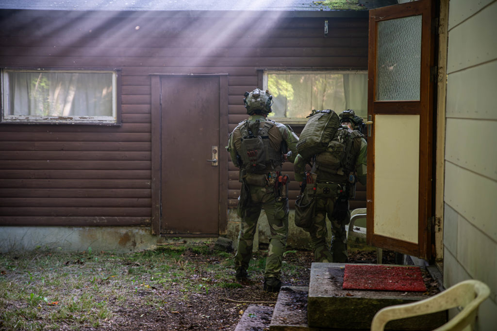 Soldater i militärkläder fotograferade bakifrån vid husvägg i motljus.