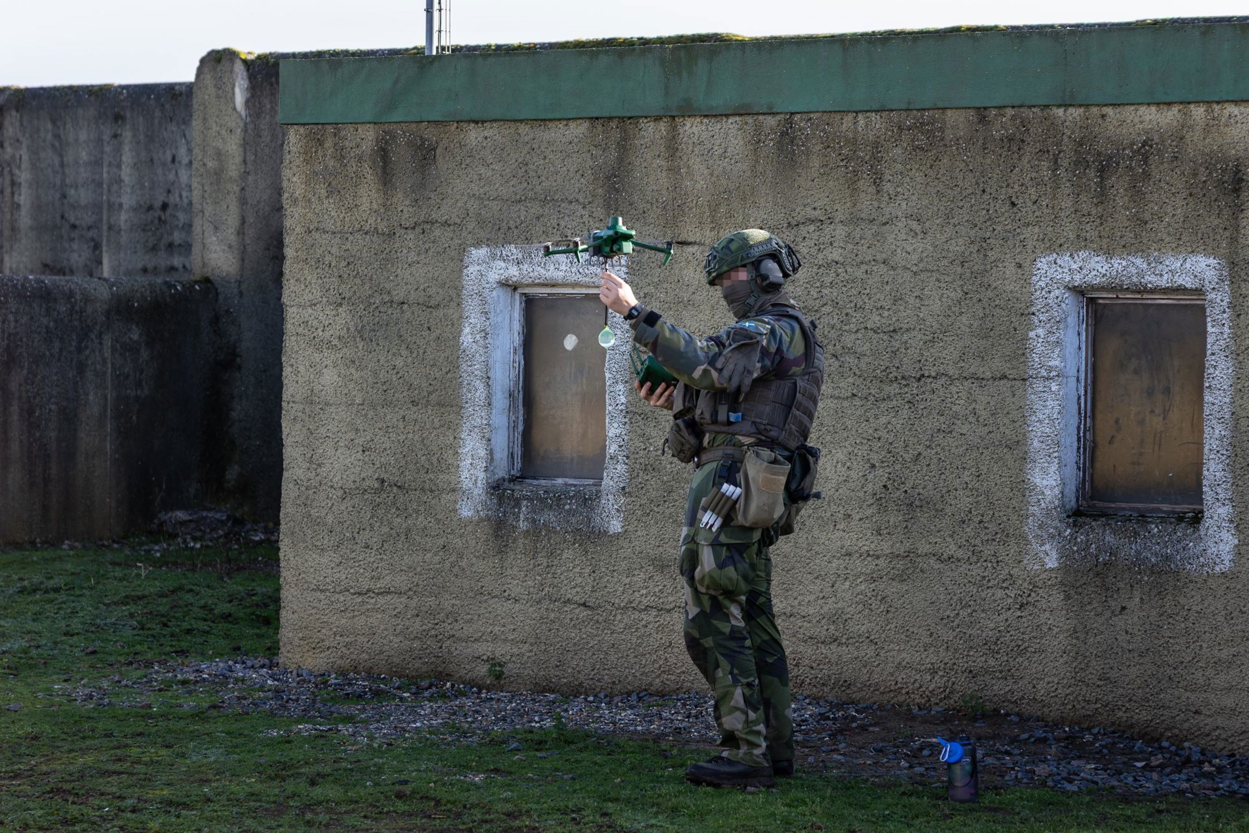 En grönklädd soldat står framför ett ljusgrönt stenhus. Soldaten är maskerad och håller i en drönare.