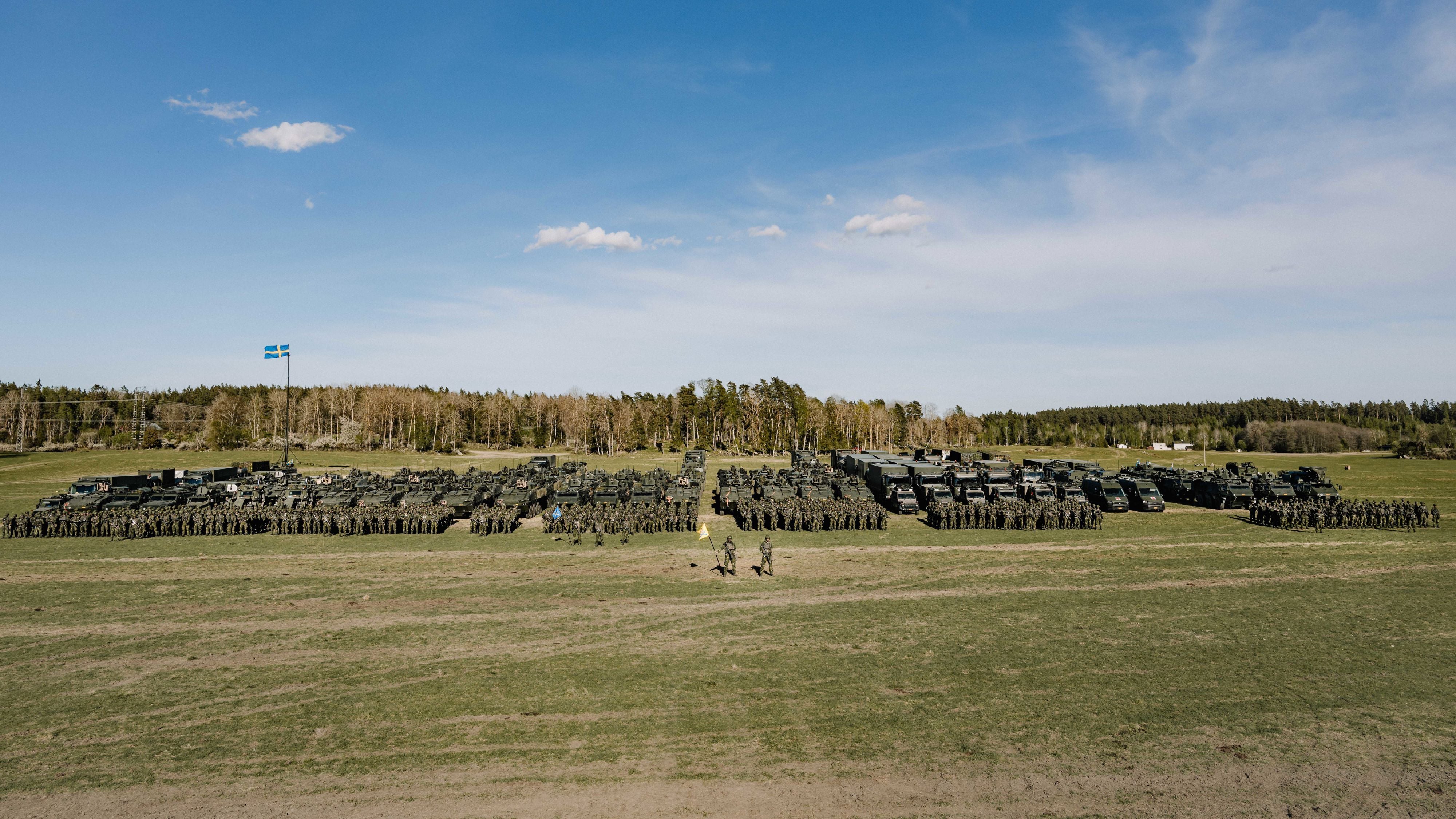 En hel brigad står uppställda på grön mark framför fordon. I bakgrunden syns träd