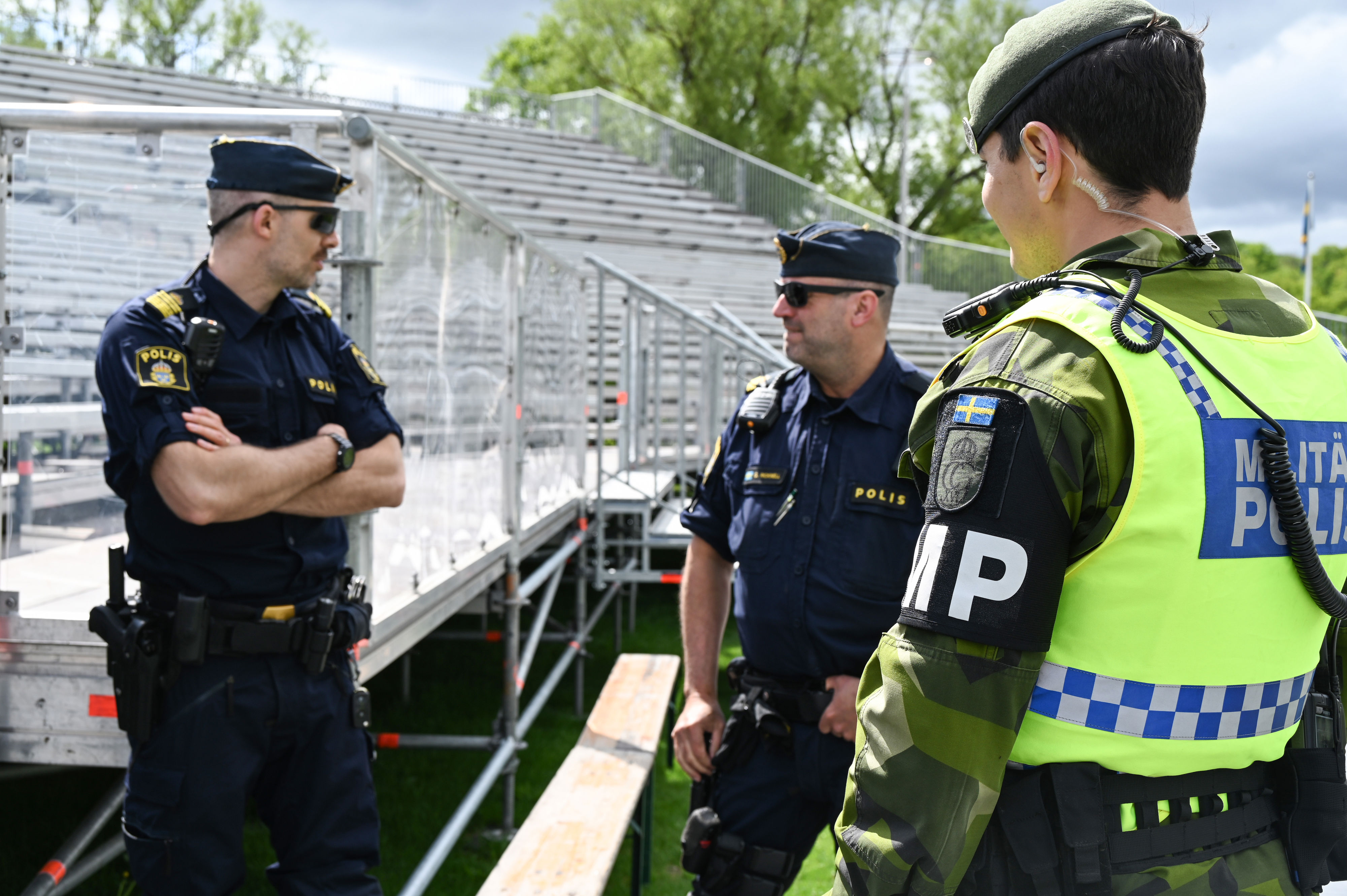 En militärpolis står och pratar med två poliser. I bakgrunden syns en läktare.