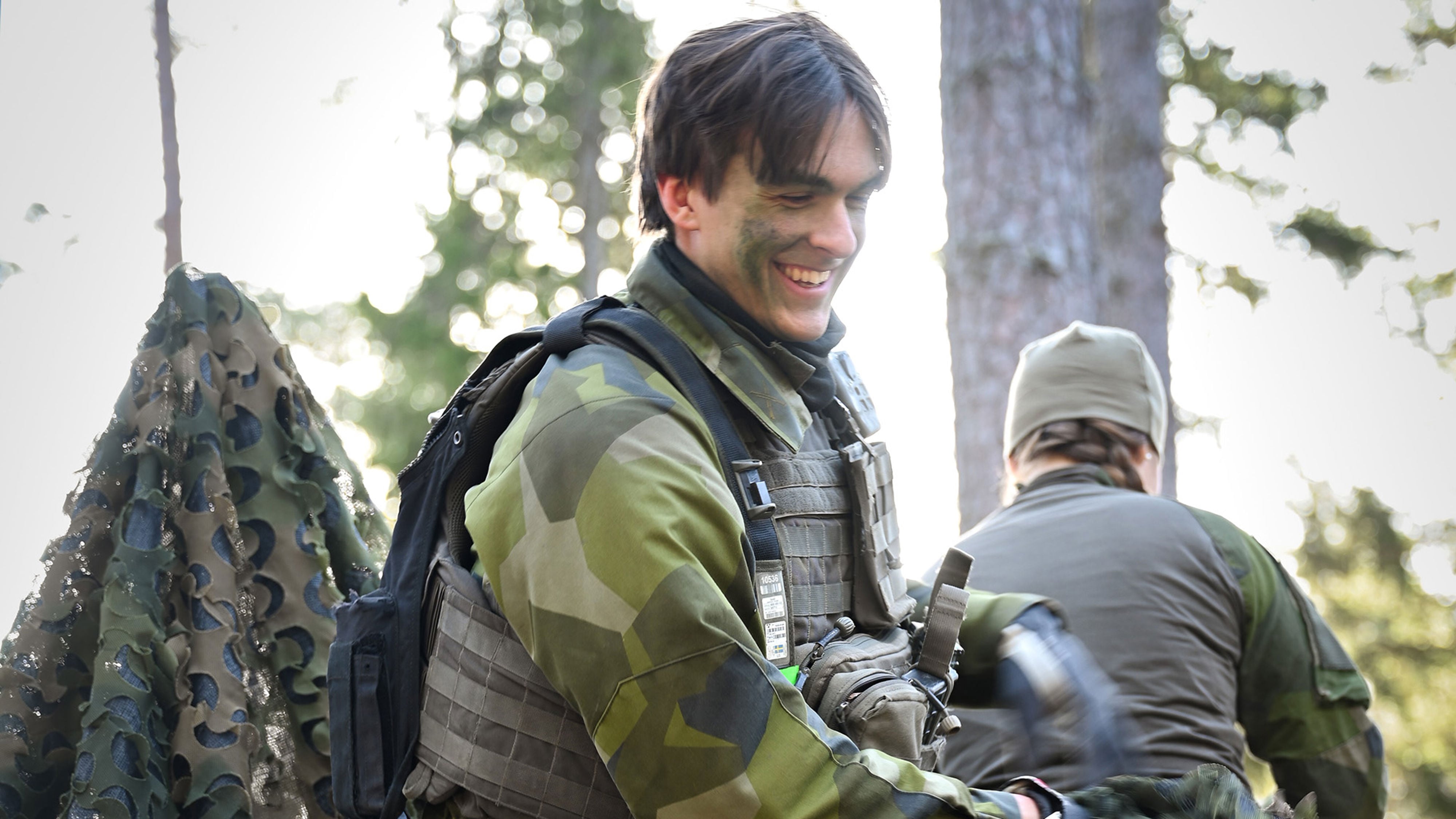 En leende soldat reser tält i skogen och ryggen på en annan soldat syns i bakgrunden.