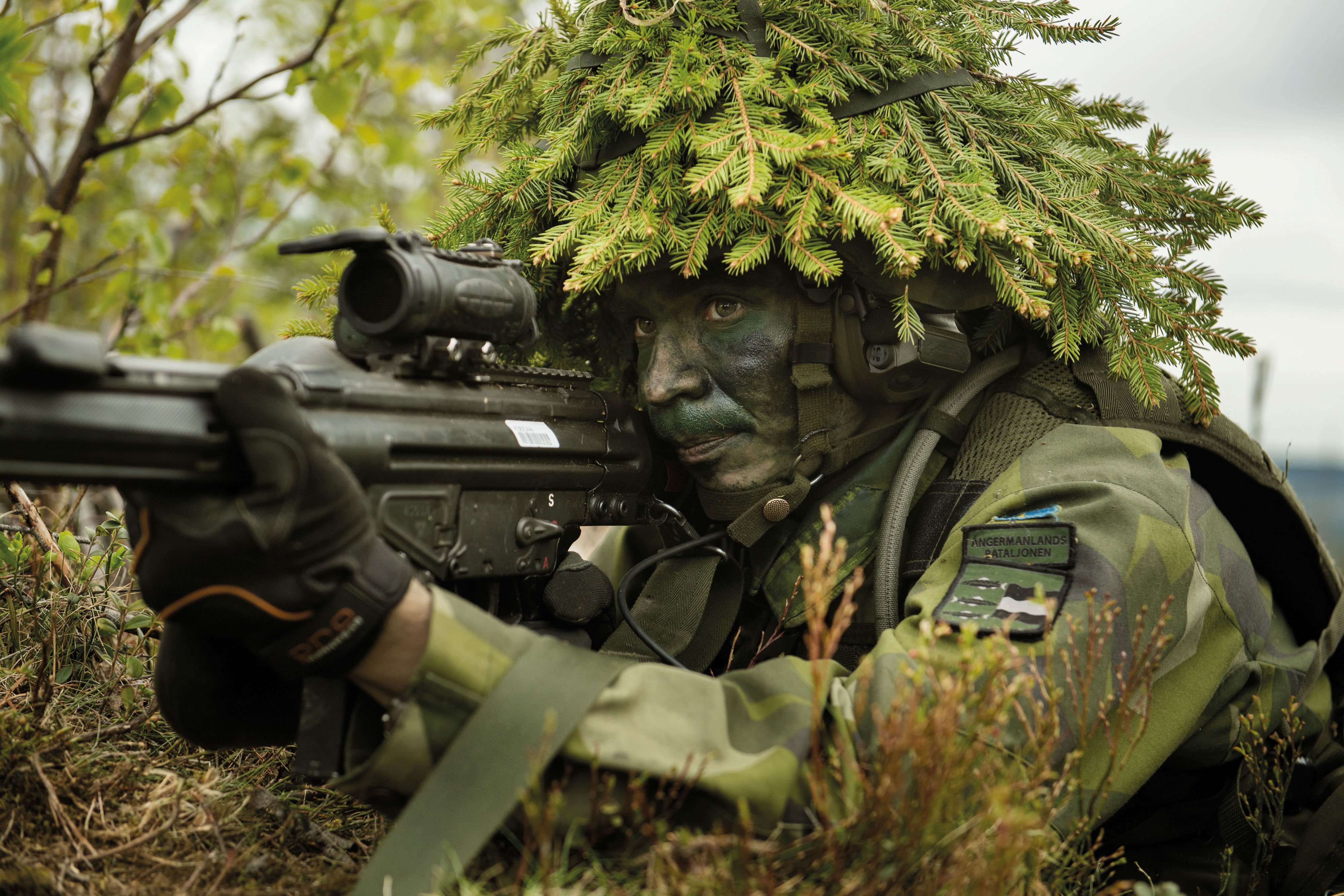 Närbild på hemvärnssoldat iklädd grön uniform och stridshjälm klädd med granris siktar med ett svart vapen.