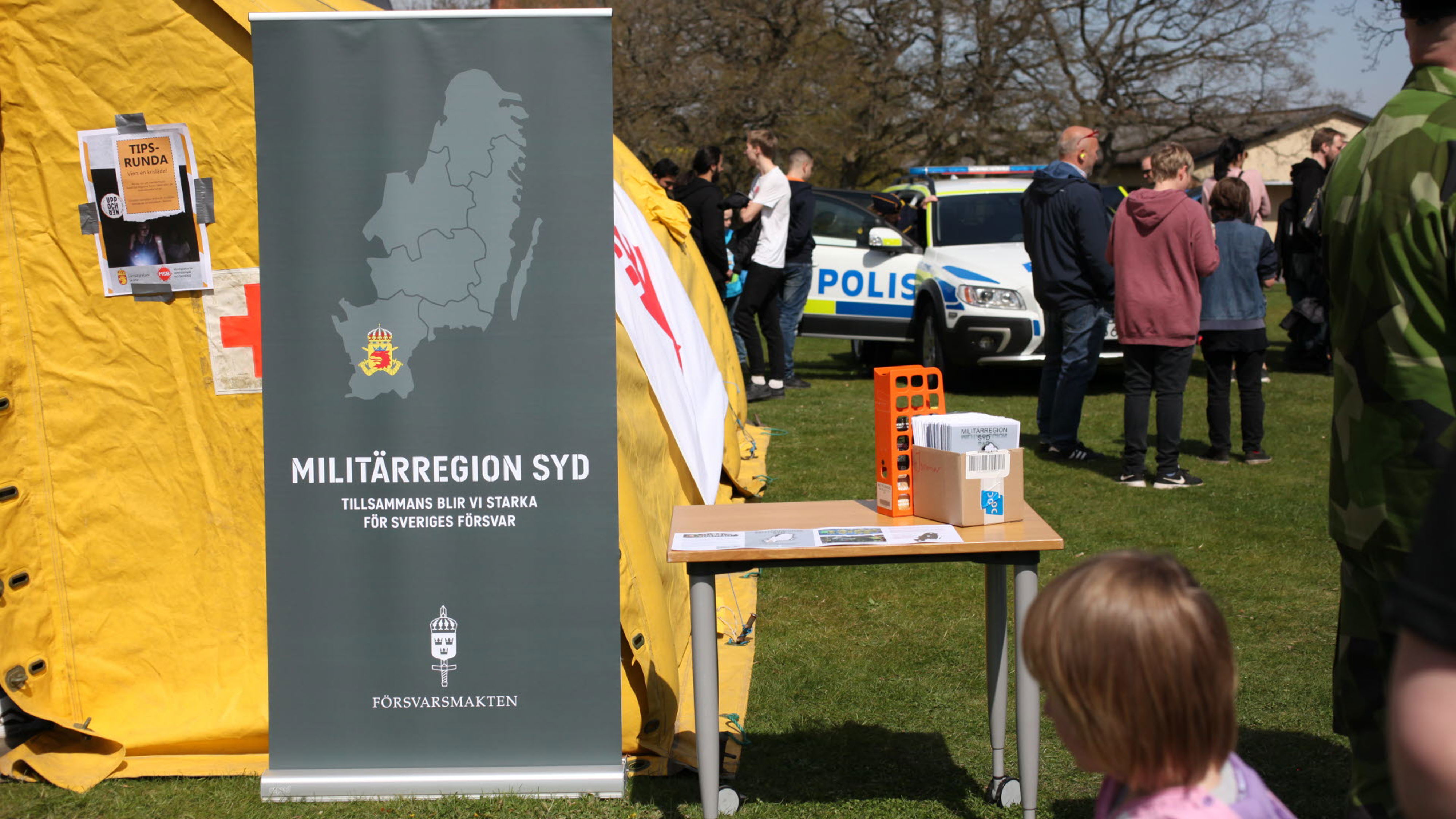 Ett event, ett bord och en rollup, besökare och en polisbil i bakgrunden.