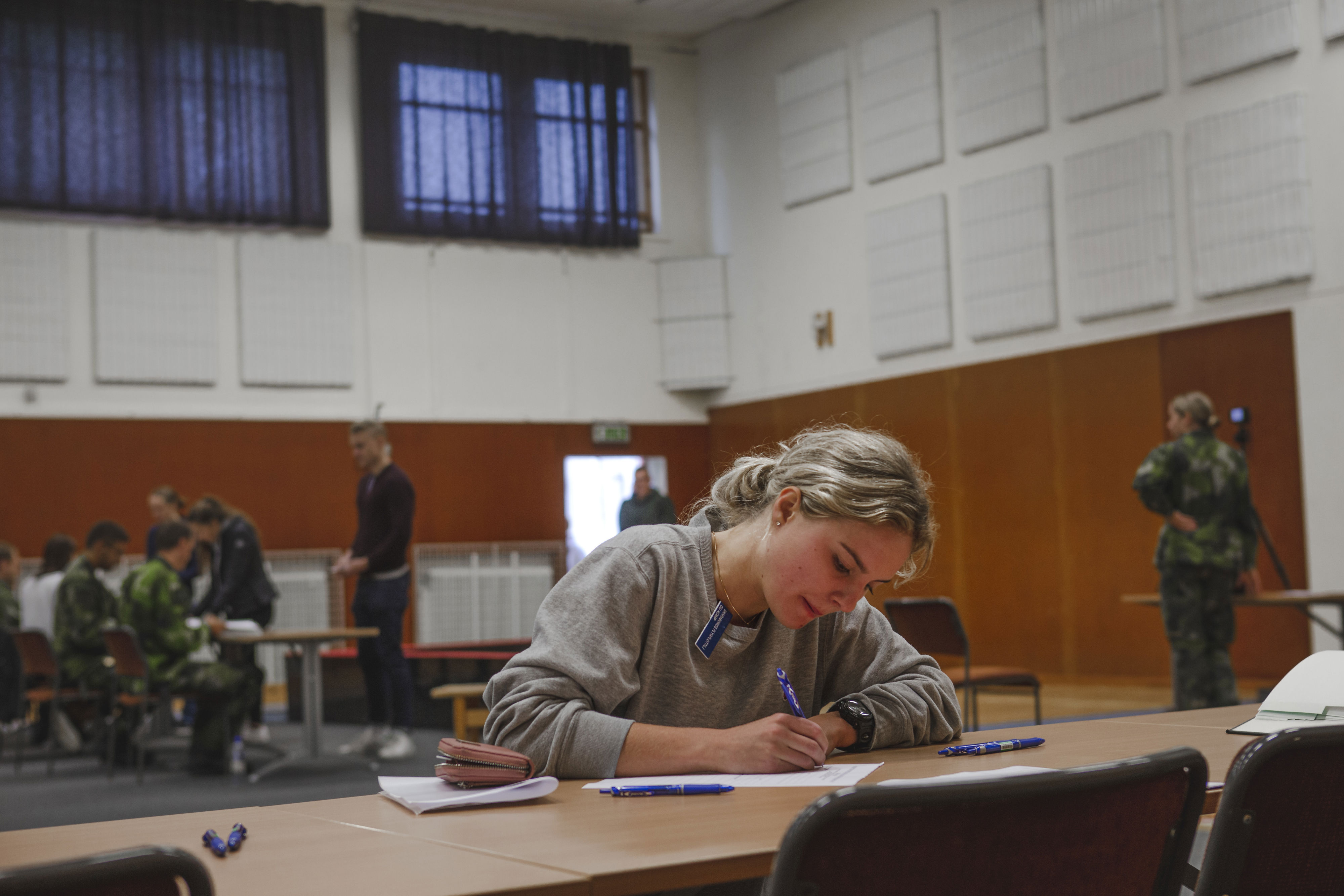 En tjej skriver på ett papper i en sal med människor i bakgrunden.