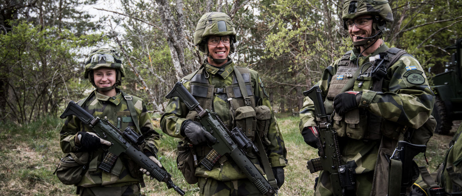 Tre militära personer står i skogen och ler mot varandra.