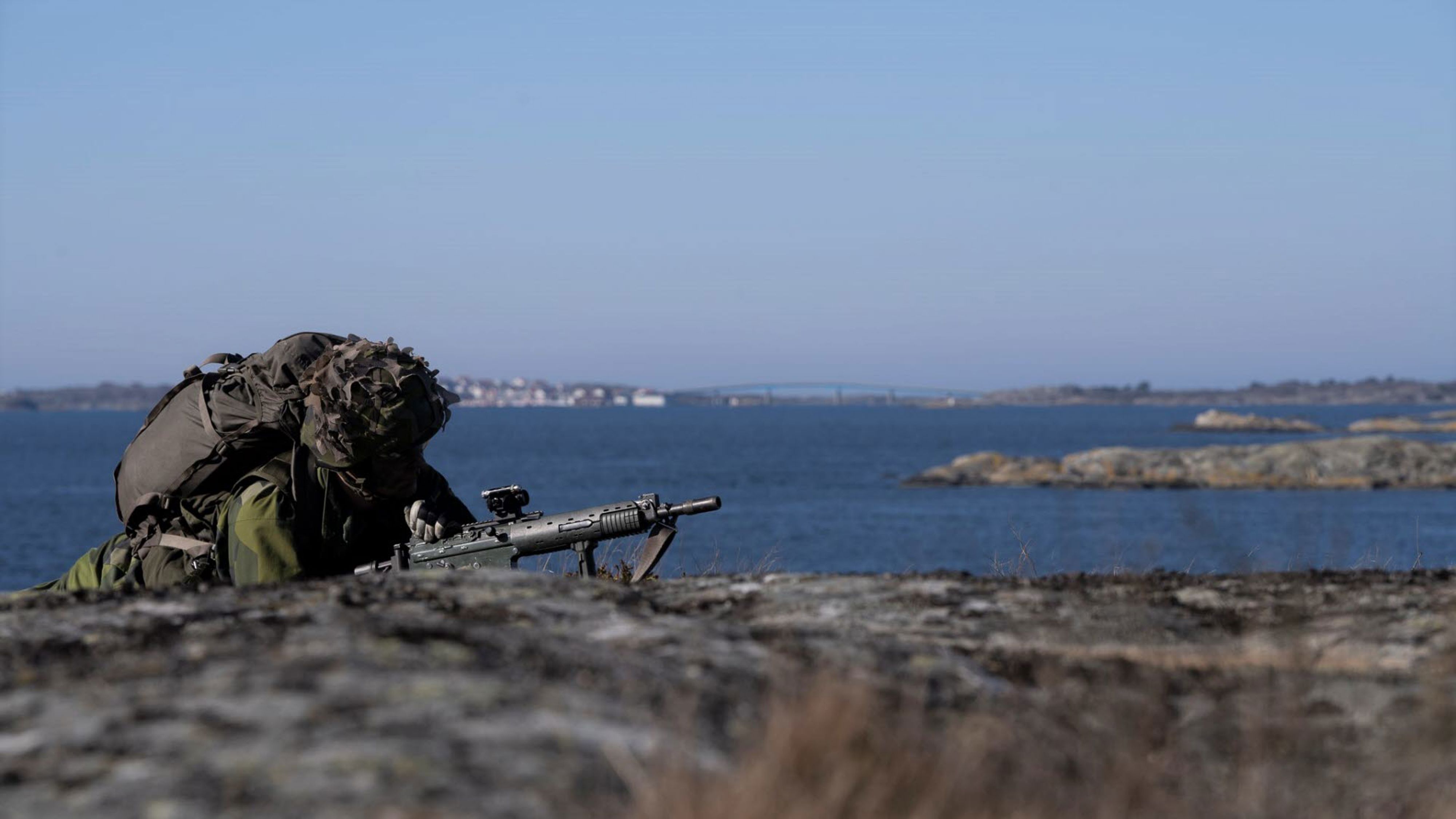 Soldat som ligger med gevär på en klippa nära vattnet.